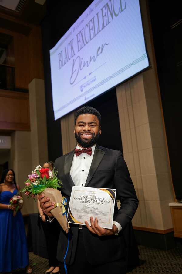 ND Law 3L Jakim Aaron standing in a black suit with red bow tie holding flowers and a certificate for the Black Excellence Award