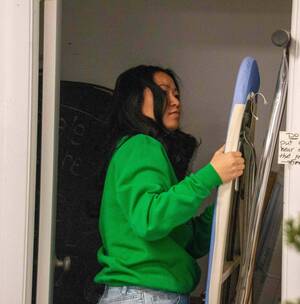 A person wearing a green sweatshirt and jeans places an ironing board into a narrow closet. A handwritten sign on the closet door reads, "Do NOT put anything near extinguisher that reduces access—Fire Hazard." A partially obscured fire extinguisher is visible near the ironing board.