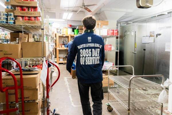 A student wearing a blue "Martin Luther King Jr. 2025 Day of Service" t-shirt walks down the center aisle of a food pantry stocked with shelves full of canned goods and boxes.  Metal rolling carts flank the aisle.