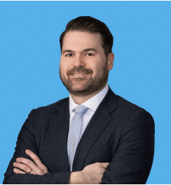 Matthew Lamb, a man with short brown hair and a beard smiles at the camera against a bright blue background. He wears a dark suit jacket, light blue tie, and has his arms crossed.