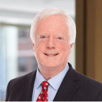 A headshot of Michael Scanlon Jr., a smiling man with white hair, wearing a dark suit, light blue shirt, and red patterned tie. He stands before a blurred office background with windows.