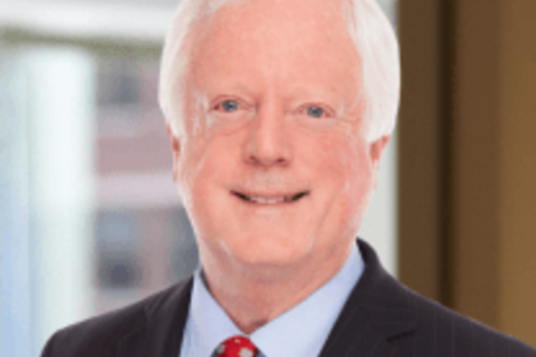 A headshot of Michael Scanlon Jr., a smiling man with white hair, wearing a dark suit, light blue shirt, and red patterned tie. He stands before a blurred office background with windows.