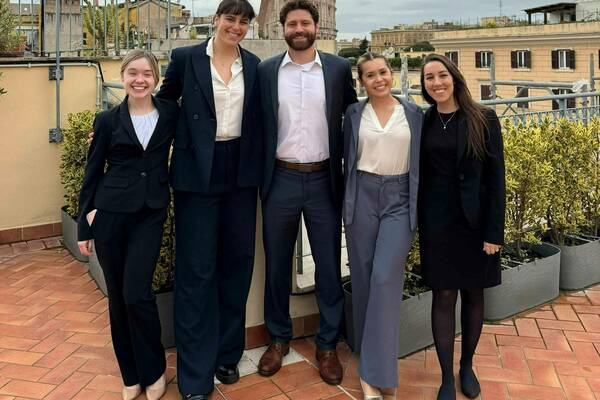 Four students and Professor Jacquline Carney in business attire pose on a rooftop in Rome, Italy, with the Colosseum visible in the background under a cloudy sky.
