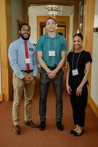 Three people wearing name tags stand in a hallway with light wood accents and reddish-brown patterned carpeting.  They are smiling and have their hands clasped in front of them.  The person on the left wears a light blue, long-sleeved shirt, khaki pants, and brown dress shoes.  The center person wears a teal polo shirt, gray pants, and black shoes.  The person on the right wears a black jumpsuit and black flats.