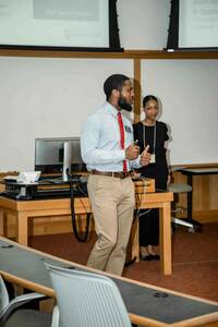 A man wearing a light blue shirt, khaki pants, and a red tie gestures while presenting in a University of Notre Dame classroom. A woman wearing a black dress stands behind him. Projected on screens are University branding and presentation slides.