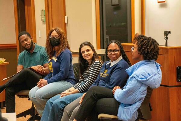Five people sit in chairs in a room. They appear to be listening to a presentation. One person wears a Notre Dame sweatshirt and another wears a Saint Mary's College sweatshirt. A webcam is positioned on a wooden cabinet.
