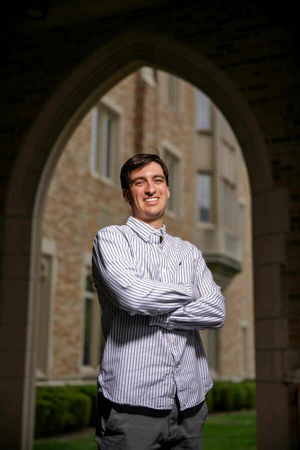 A smiling man in a blue and white striped shirt and gray pants stands with his arms crossed under a stone archway on the Notre Dame campus.