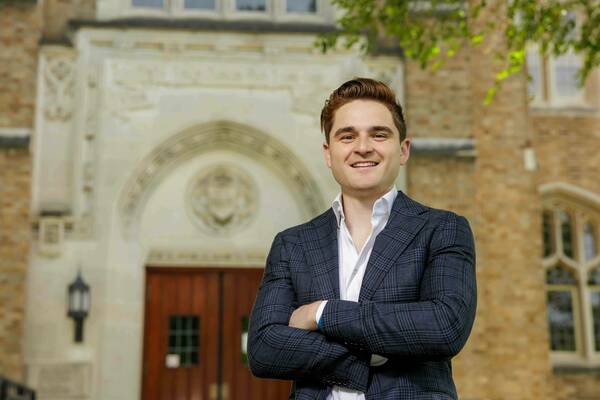 Paul Cuccinelli, a smiling man with arms crossed, wearing a blue plaid sport coat and white shirt, stands in front of the Main Building at the University of Notre Dame.