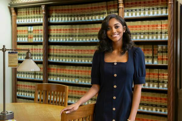 Kaitlyn Bowe, a smiling student in a navy blue dress stands next to a wooden chair and table in front of bookshelves filled with legal volumes.
