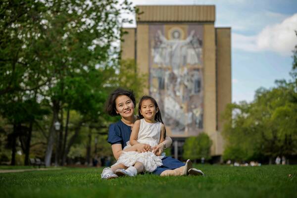 A woman and young girl sit smiling on the grass in front of Hesburgh Library at the University of Notre Dame.  The woman wears a navy dress and the child a white ruffled dress.  Touchdown Jesus is visible on the library facade.