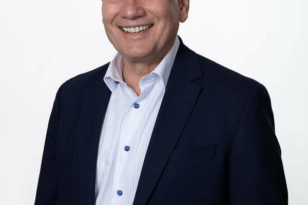 Man with light brown hair wearing a navy blue blazer and a white and light blue striped shirt smiles at the camera against a white background.