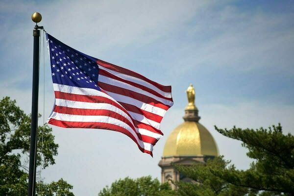 A United States flag waves in the foreground, with the Golden Dome of the Main Building at the University of Notre Dame blurred in the background.