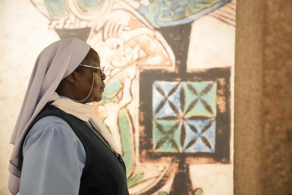 A nun in a light blue habit and white veil views a framed piece of art.