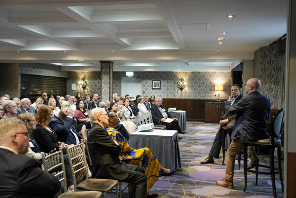 Two men converse on stage before a seated audience in a conference room. One sits and gestures while the other perches on a tall stool. The audience members wear a variety of business and business casual attire.
