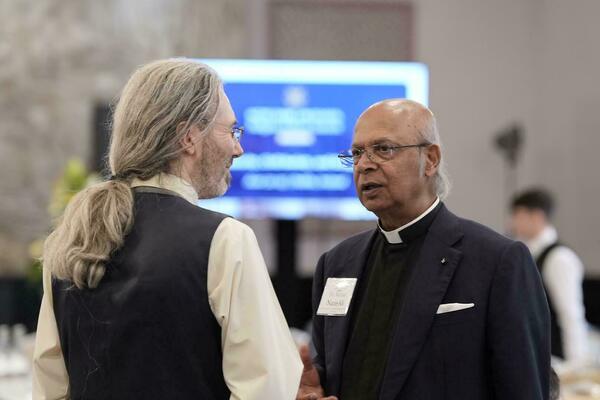 A person with long gray hair tied back speaks with Dr. Michael Nazir-Ali, who wears a clerical collar and a navy blue suit.