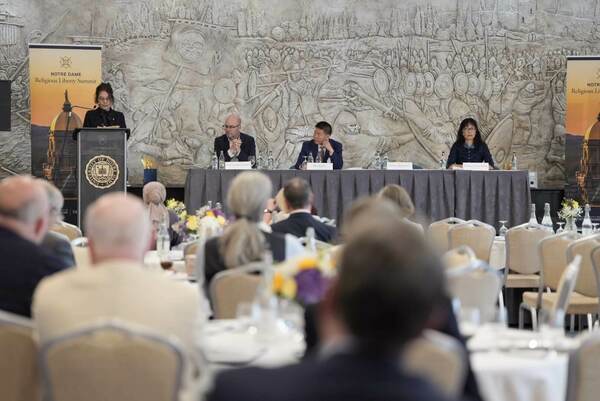 A woman speaks at a podium with a Notre Dame Religious Liberty Summit banner beside it. Three panelists sit at a table behind her. A large carved mural of a battle scene covers the wall behind them. Audience members are seated at round tables in the foreground.