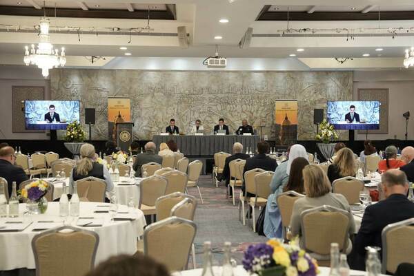 Audience members seated at round tables watch speakers at a head table, with a large relief mural behind them and Notre Dame Religious Liberty Summit banners and screens displaying a remote speaker.