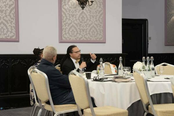 A person with long locs, wearing glasses, a black suit, and a gold bow tie, speaks into a microphone while gesturing with their hand at a round table during a meeting.  Other attendees sit around the table, listening.