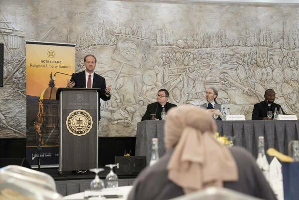 A man in a suit and tie gestures while speaking at a podium with a Notre Dame logo. Behind him, a large relief depicts a historical battle scene. Three men are seated at a table in the background. A person wearing a light brown head covering is slightly out of focus in the foreground. A banner for the Notre Dame Religious Liberty Summit is visible to the left of the podium.