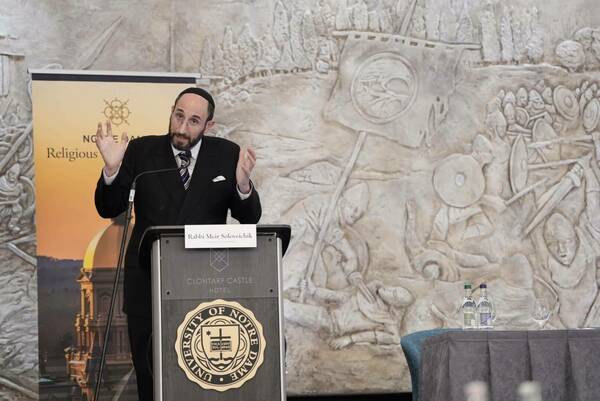 Rabbi Meir Soloveichik gestures while speaking at a podium bearing the University of Notre Dame seal at the Clontarf Castle Hotel. A large relief depicting a medieval battle scene is visible on the wall behind him.