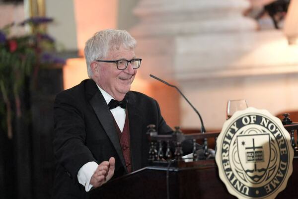 A gray-haired man in a tuxedo and glasses smiles while gesturing with his hand at a podium bearing the University of Notre Dame seal.