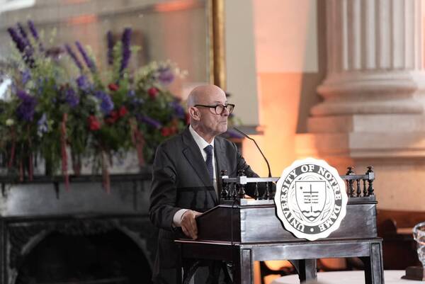 A man in a suit and tie speaks at a podium bearing the University of Notre Dame seal. A large floral arrangement is visible behind him.