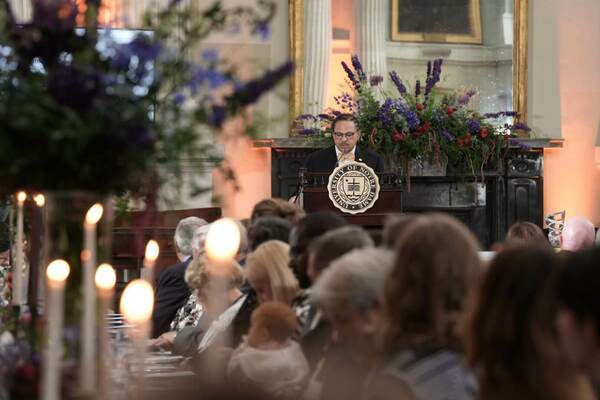 A speaker stands at a podium bearing the University of Notre Dame seal, addressing an audience seated at tables with lit candles and floral centerpieces.