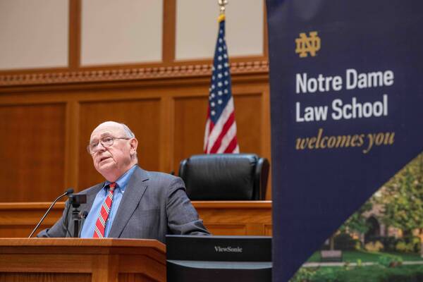 A man in a gray suit and red-striped tie speaks at a wooden podium. An American flag and a Notre Dame Law School banner stand behind him.