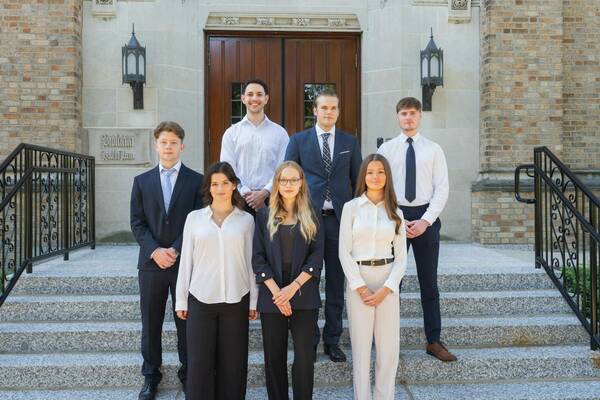 Seven students in business attire pose on the steps in front of Biolchini Hall of Law.