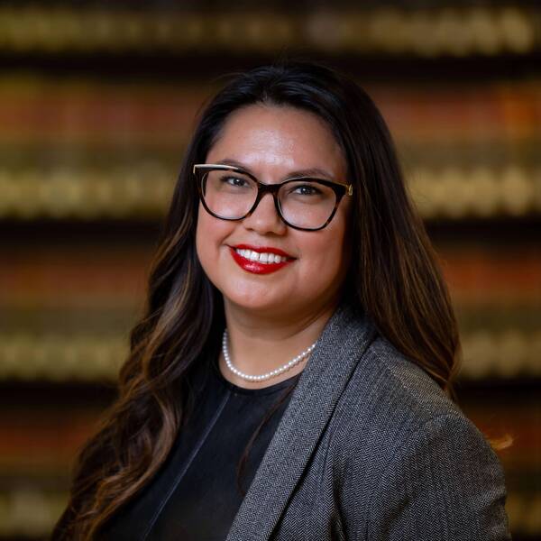 A smiling woman with long, dark, wavy hair, wearing patterned glasses with gold accents, bright red lipstick, a pearl necklace, and a gray blazer.