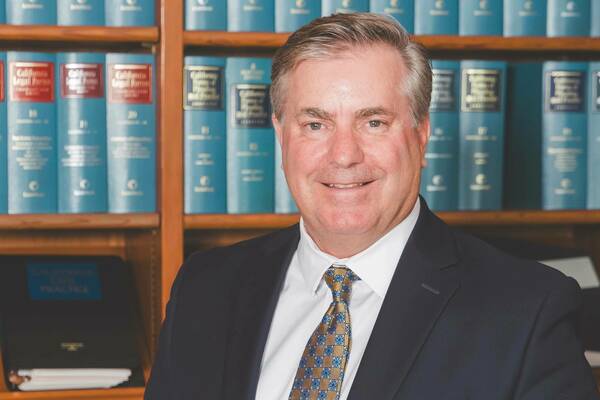 A smiling man with graying hair, wearing a dark suit, white shirt, and gold and blue patterned tie, stands in front of a bookshelf filled with blue legal books, some labeled "California Legal Forms."