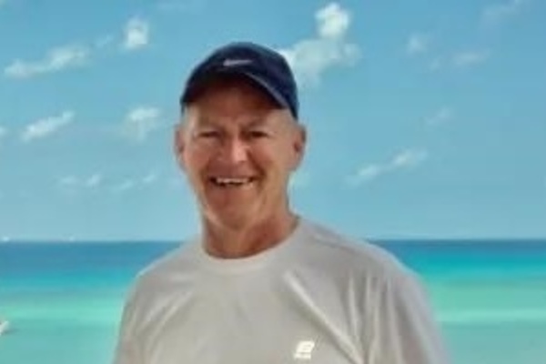 Man wearing a baseball cap and long-sleeve shirt smiles while standing outdoors near a turquoise ocean, with boats visible in the background under a clear sky.