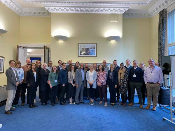 A diverse group of approximately 30 professionals smile while standing in a room with light yellow walls, ornate white molding, and blue patterned carpet. Many wear lanyards.