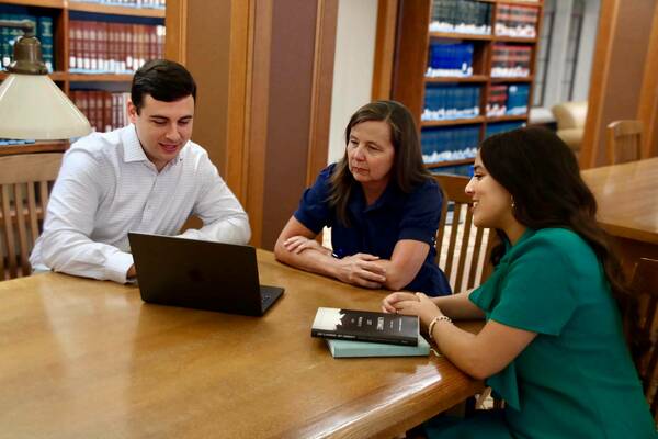 Three people collaborate at a wooden library table. A man in a white shirt looks at a laptop. A woman in a navy dress listens as a woman in a teal top smiles and speaks. Bookshelves line the background.