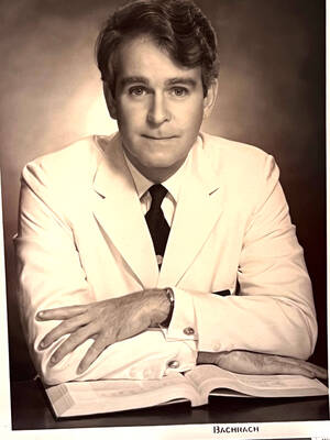 Sepia-toned portrait of a man with wavy brown hair, wearing a light suit jacket, white shirt, and dark tie. He looks directly at the camera with a composed expression, his hands resting on an open book.