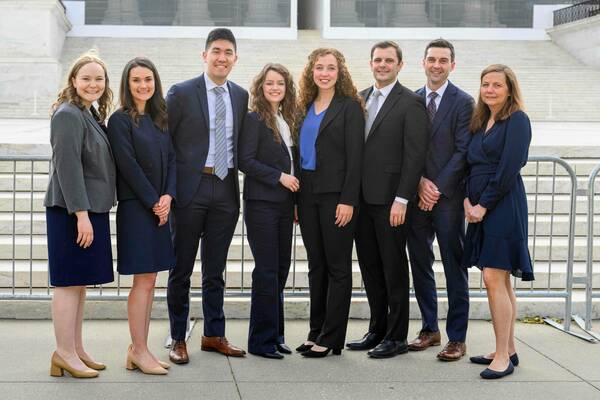 Eight smiling individuals in dark professional attire stand on a paved surface before a stately, light-colored stone building with wide steps and a metal barrier.