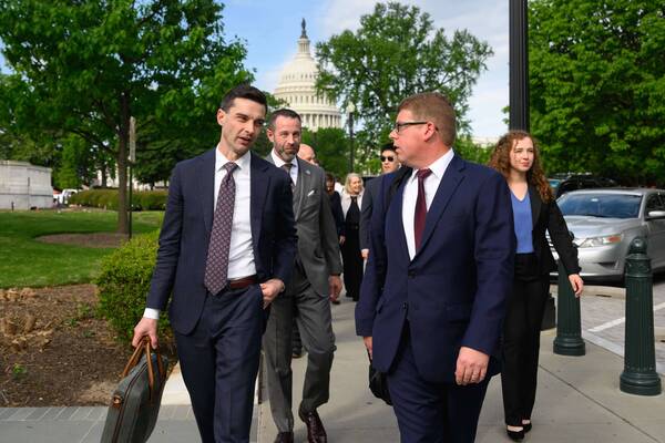 Two men in dark blue suits, one speaking and carrying a briefcase, walk on a paved path alongside a woman in a black blazer. The U.S. Capitol Building dome is visible behind green trees.
