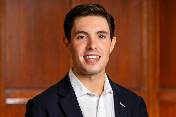 Young man with short dark hair smiling, wearing a navy suit jacket and white shirt against a warm brown paneled background.