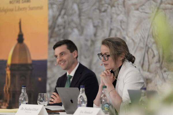 Dr. Ewelina Ochab, in a white blazer and black glasses, sits with hands clasped, speaking at the Notre Dame Religious Liberty Summit. A man in a dark suit and green tie smiles next to her. The Gold Dome appears on a banner behind them.