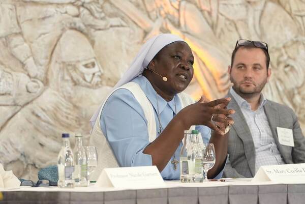 Sr. Mary Cecilia D., a nun in a light blue habit and white veil, speaks animatedly into a lavalier microphone, a crucifix on her necklace. She gestures with her hands as Dr. Michael Holland, a man in a gray suit, sits behind her. A carved stone wall is in the background.
