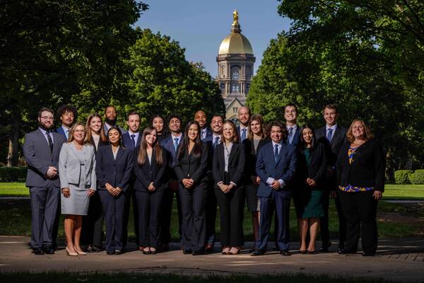 A diverse group of 25 smiling individuals, dressed in dark business suits and dresses, stands on the Notre Dame campus with the Golden Dome of the Main Building and green trees under a blue sky.