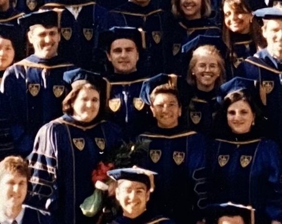 Smiling Notre Dame graduates in dark blue academic regalia with gold trim and crests. A woman in the foreground holds a bouquet of red and green flowers.