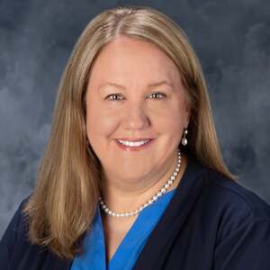 Smiling woman with long blonde hair, a pearl necklace and earrings, wearing a blue top and navy jacket, against a mottled dark gray background.