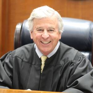 A smiling man with white hair wears a black judge's robe, white shirt, and yellow patterned tie, seated in a dark leather chair against a wooden wall.