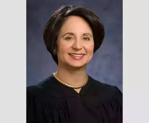 Sonia Sotomayor smiles, wearing a black judicial robe, a gold choker necklace, and small gold earrings against a mottled blue background.