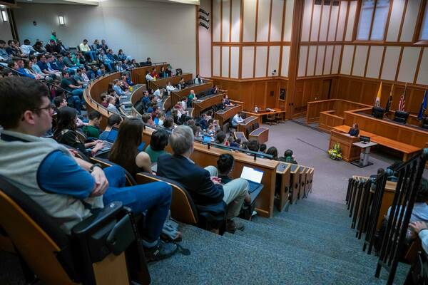 A tiered lecture hall with wood panels and light walls at the University of Notre Dame is full of students. A woman in a dark blazer speaks from a wooden podium, with US, Vatican, and Indiana flags, and a floral arrangement visible behind her.