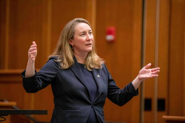A blonde woman in a navy blazer, wearing a Golden Dome pin, speaks animatedly, gesturing with both hands. She stands in a wood-paneled lecture hall.