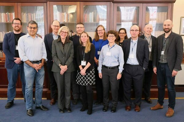 Twelve people, seven men and five women, stand and smile for a photo in front of dark wood bookshelves. Most wear lanyards with ID badges.