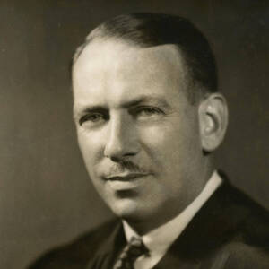 Sepia-toned headshot of a man with neatly combed dark hair and a mustache. He wears a suit, white shirt, and patterned tie, looking to the side with a neutral expression.