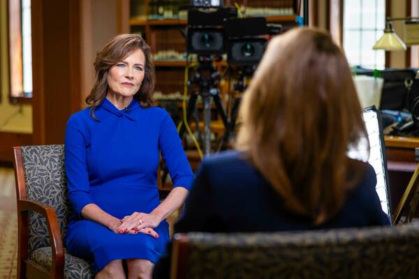 Amy Coney Barrett, in a royal blue dress, sits in a patterned chair, looking thoughtfully at an unseen interviewer. Her hands are clasped, showing red nails. A blurred library with video recording equipment is in the background.